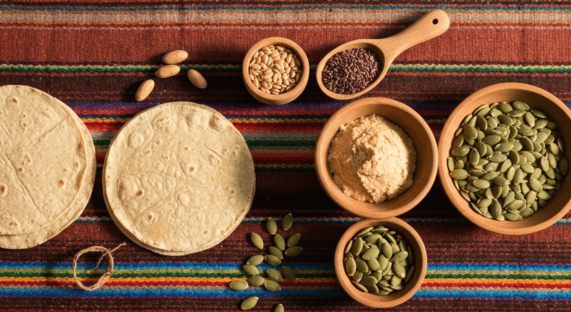 Composición tipo flat lay con ingredientes prehispánicos ricos en proteína: frijoles negros, amaranto, semillas de chía, pepitas de calabaza, tortillas de maíz y un cuenco con masa. Fondo de tela artesanal mexicana, colores terrosos y cálidos. Iluminación natural suave, foco en textura y autenticidad, estilo fotográfico naturalista.